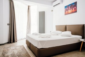 A modern hotel room with a large bed, a bedside table, a wall-mounted air conditioner, and a painting of palm trees.
