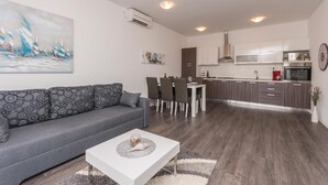 Un salon moderne avec un canapé gris, une table basse blanche et un coin repas comprenant une table et des chaises blanches. La cuisine est équipée d’appareils électroménagers en acier inoxydable et de meubles de rangement en bois.