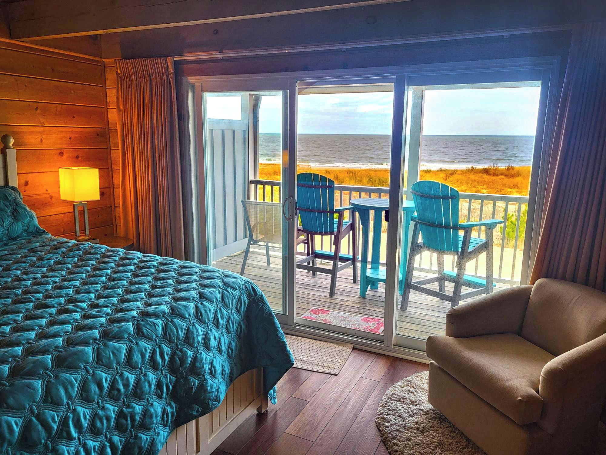 A room with a bed, a chair, and a view of the beach through a sliding door.