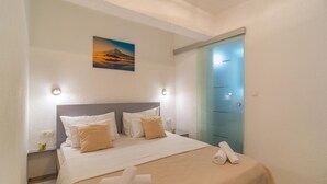 Een net opgemaakt bed met wit en beige beddengoed, een schilderij van een berg en een blauwe deur.