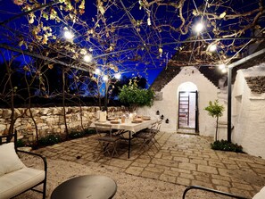 An outdoor patio with a stone wall, a table set for dining, and string lights overhead.