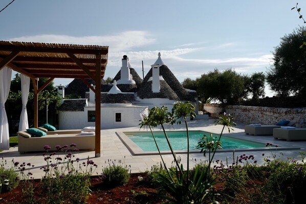 A pool area with a pergola, outdoor seating, and a stone building with conical roofs in the background.