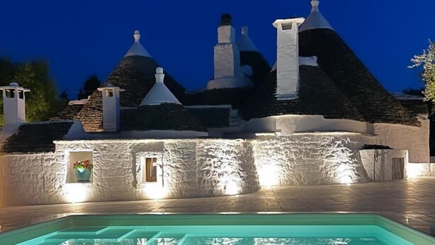 A swimming pool with a stone wall and a building with multiple chimneys in the background. - Trullo Valle d'Itria (Martina Franca)