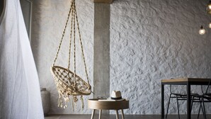 Ein Zimmer mit strukturierter Wand, einem hängenden Makramee-Stuhl, einem runden Holztisch mit Hut und Brille sowie einem schwarzen Metallstuhl.