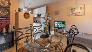 Een moderne woonkamer met een glazen salontafel, zwarte metalen stoelen, een open haard, een televisie en een keuken op de achtergrond.