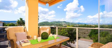 A balcony with wicker furniture, a table, and a view of green hills and a clear sky.