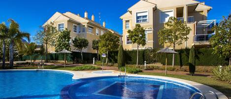 A swimming pool with a clear blue water, surrounded by a well-maintained garden and a two-story building with balconies.