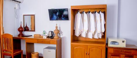 A room with a wooden desk, a chair, a small refrigerator, a television, a mirror, a cabinet with hanging robes, and a small cabinet with a radio.