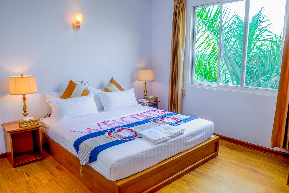 A neatly made bed with a floral bedspread, two bedside lamps, and a window with curtains.