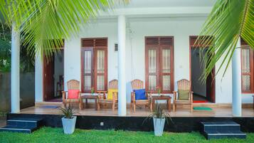 A covered porch with wooden chairs and tables, surrounded by potted plants and palm trees.