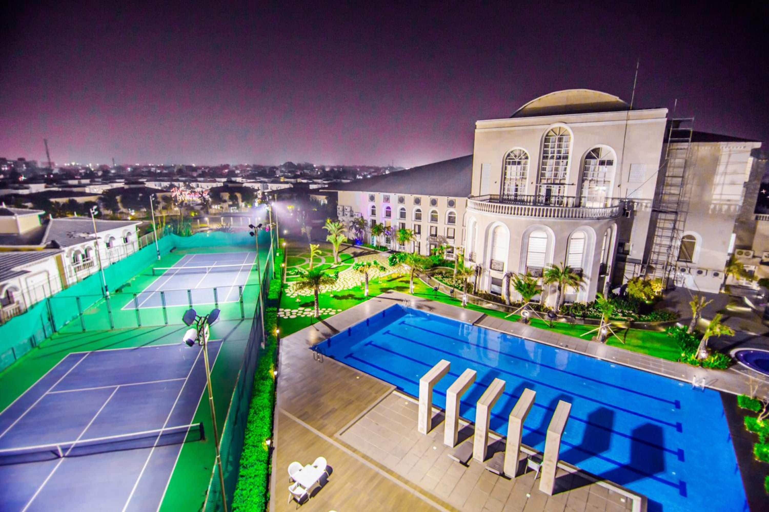 Un espace piscine aménagé sur le toit, avec des courts de tennis et une piscine, le tout entouré de verdure et bordé d’un bâtiment aux fenêtres en arc de cercle.