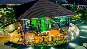 Un espacio exterior moderno para comer, con varios niveles, paredes de vidrio, iluminación ambiental verde y una zona de estar central.