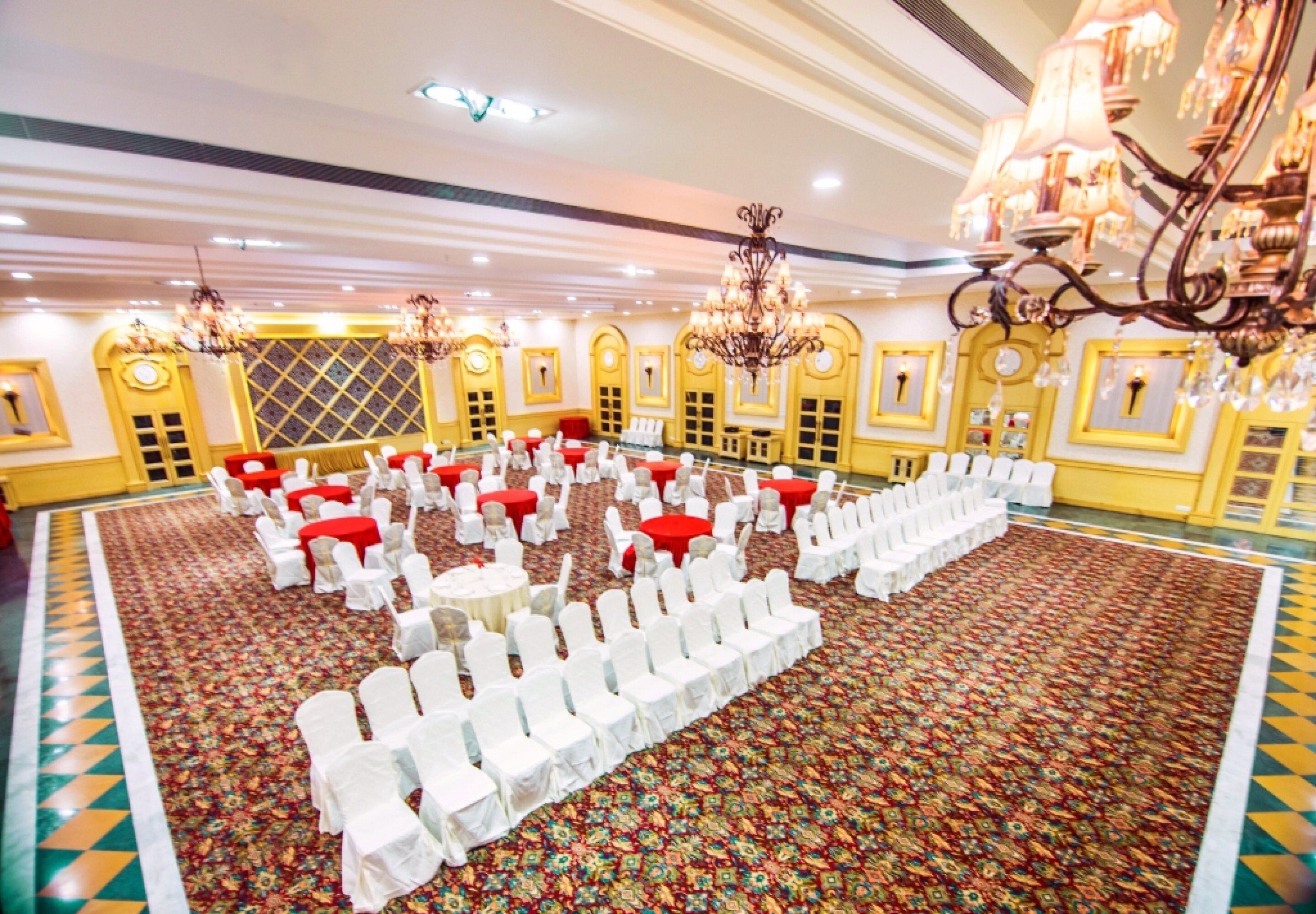 A banquet hall with rows of white chairs and tables, ornate chandeliers, and patterned carpet.