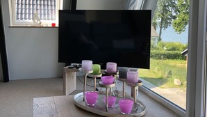 Ein Zimmer mit großem Fenster, einem Flachbildfernseher und einem Holztisch mit rosa Kerzenhaltern.