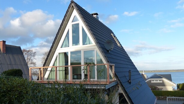 Ein modernes Haus mit einem geneigten Dach, großen Fenstern und einem Balkon mit Blick auf ein Gewässer.