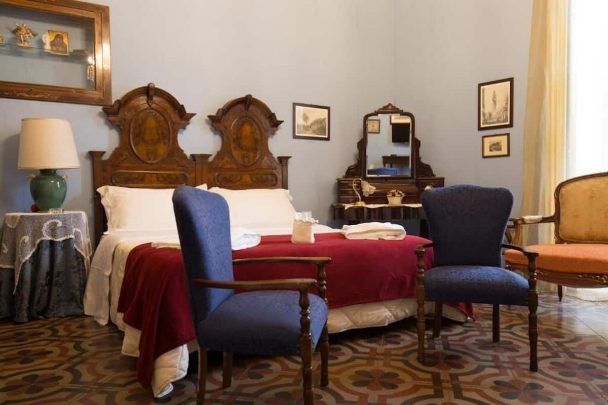 A hotel room with a wooden bed, two blue armchairs, a wooden dresser, and a patterned floor.