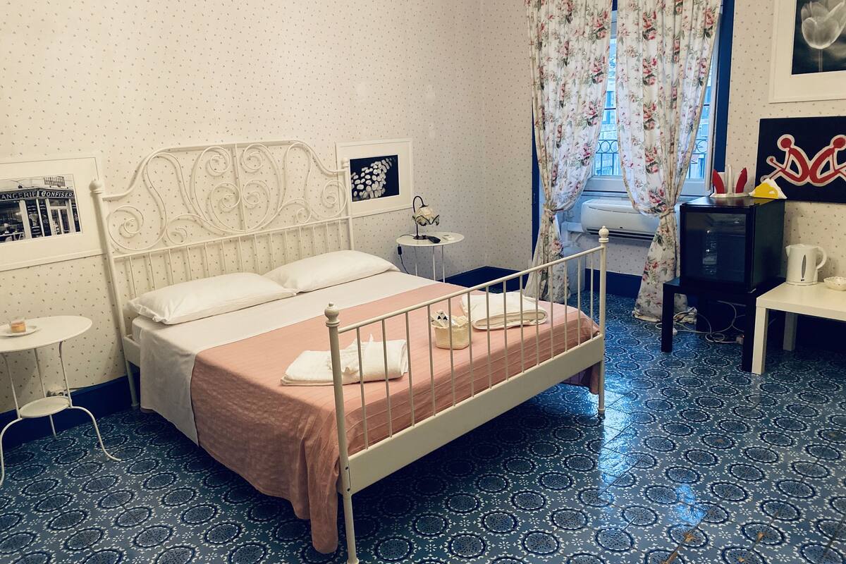 A bedroom with a bed, a nightstand, a small table, a television, and a window with curtains.