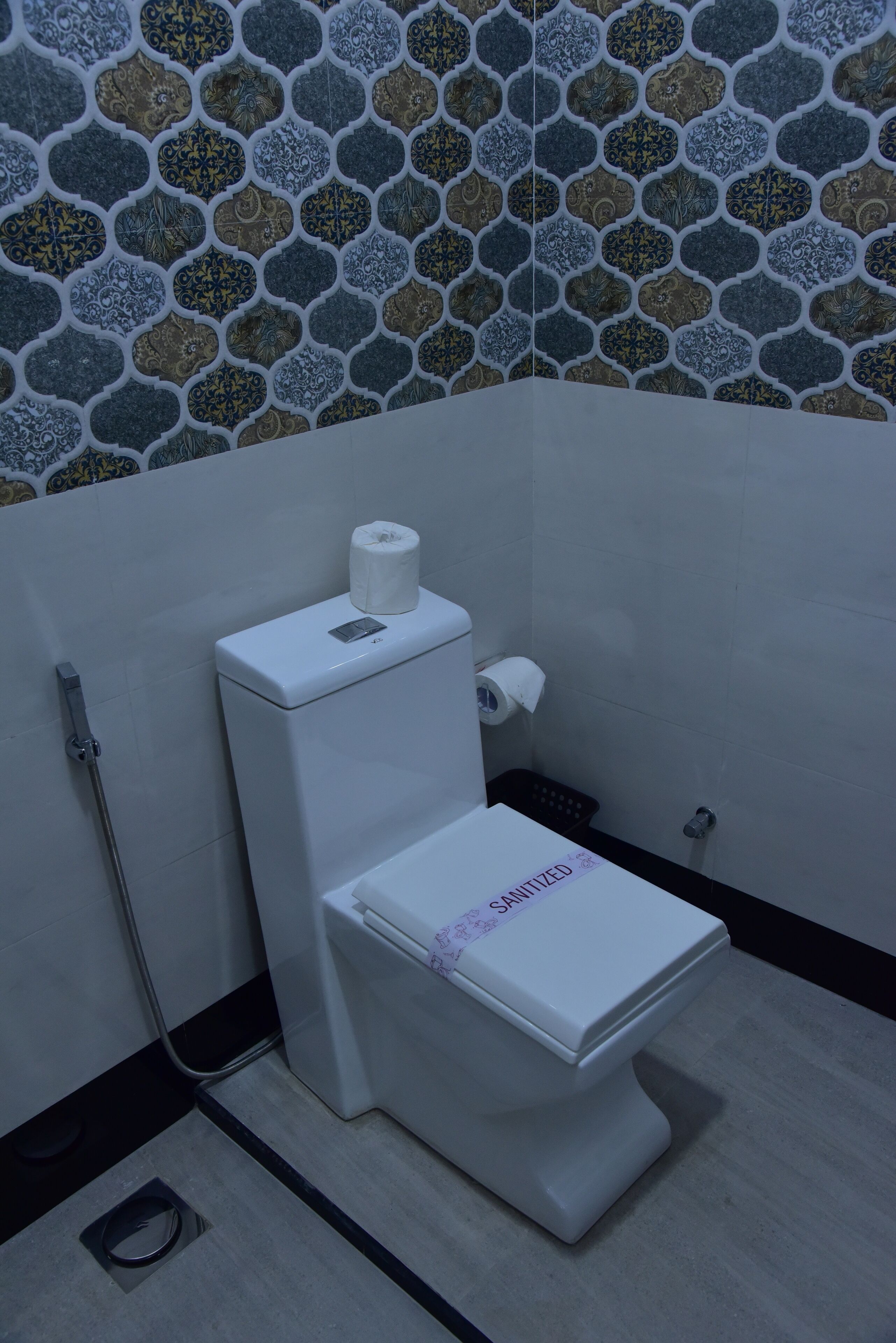 A toilet with a white seat and lid, a roll of toilet paper on the wall, and a metal hose on the floor.