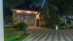 A well-lit house with a paved driveway, surrounded by greenery and palm trees.