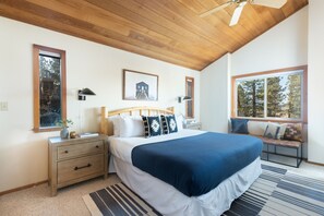 Ein Schlafzimmer mit Holzbalkendecke, einem großen Bett mit blauer Decke, einem Nachttisch, einer Bank am Fenster und einem gerahmten Bild an der Wand.