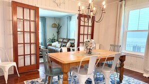 A dining area with a wooden table, white chairs, a chandelier, and a view through a window. - Modern East Beach 3BR 2BA With Pool + Walk to Beach (St Simons)