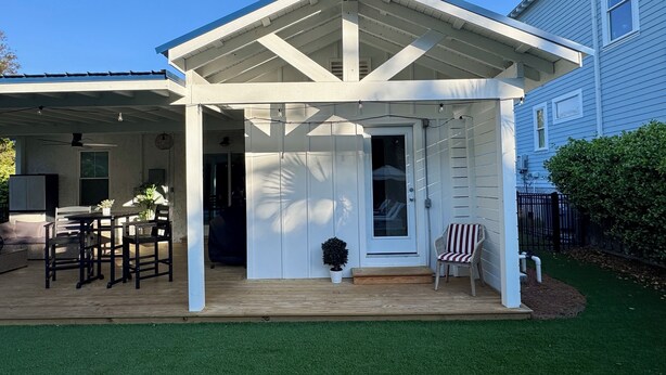 A covered patio with a wooden deck, a door, and a small seating area with a chair. - Modern East Beach 3BR 2BA With Pool + Walk to Beach (St Simons)