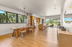 A spacious kitchen with wooden flooring, a dining area with a long table and chairs, and large windows offering a view of the outdoors.