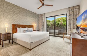 Quarto de hotel com uma cama grande, ventilador de teto, escrivaninha com televisão e vista para um jardim tropical através da porta de vidro.