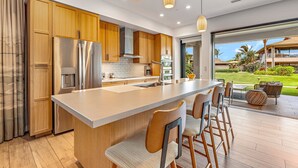 Een moderne keuken met een centraal eiland, roestvrijstalen apparatuur, houten kastjes en uitzicht op een tropisch landschap.