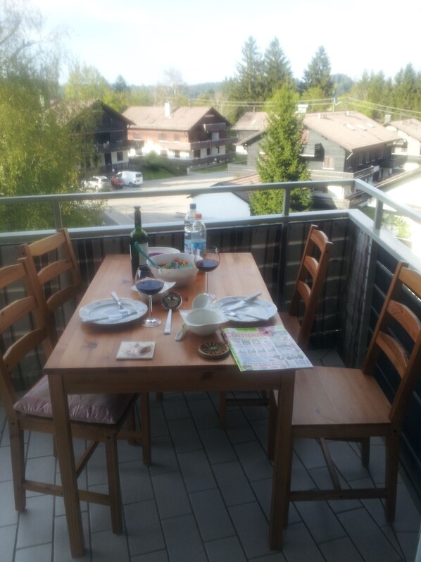 Een houten tafel gedekt voor vier personen op een balkon, met stoelen, een waterfles, een fruitschaal en een menukaart.