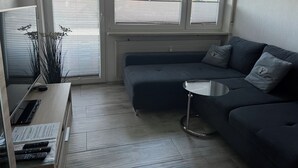 A modern living room with a dark grey sofa, a small round table, and a TV on a wooden stand. Large windows offer a view of trees and a body of water. - Apartment overlooking the Grünten, on foot to the Great Alpsee (Bühl a.Alpsee)