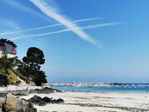 Una playa costera con un litoral rocoso, un edificio de techo inclinado y un puerto deportivo con numerosos barcos.