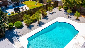 A rectangular swimming pool with a tiled edge, surrounded by a white deck. There are lounge chairs and a barbecue grill in the background.