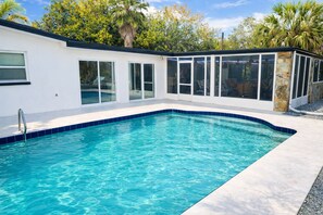 A swimming pool with clear blue water, surrounded by a white building with large windows and a stone pillar.