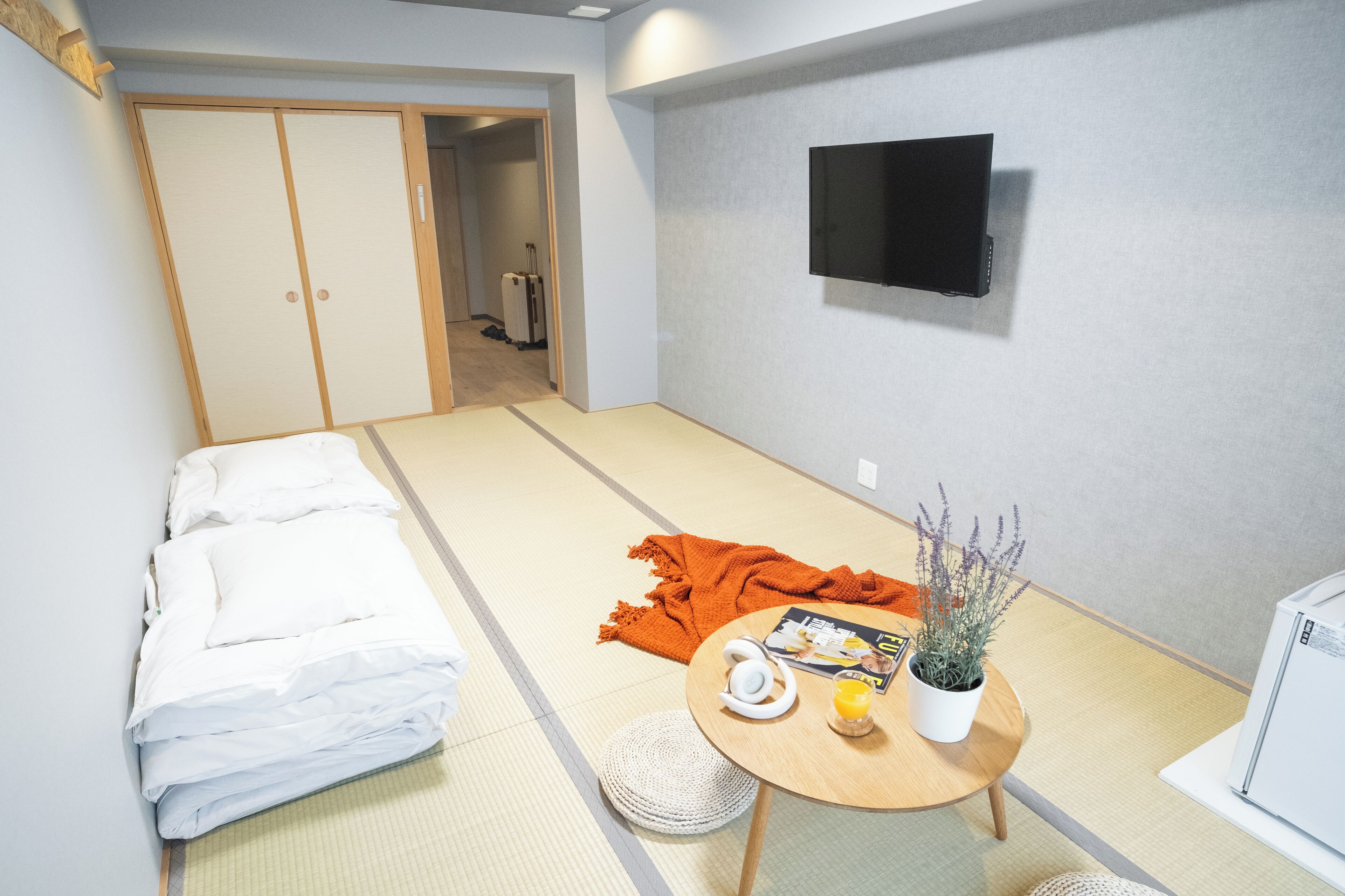 A room with tatami flooring, a bed with white bedding, a small round table with a plant, a television mounted on the wall, and a suitcase in the background.