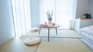 A room with tatami flooring, a round table with books and a plant, a bed with white bedding, and a window with sheer curtains. - Wayfarer Hotel Kyoto Shijo (Kyoto)