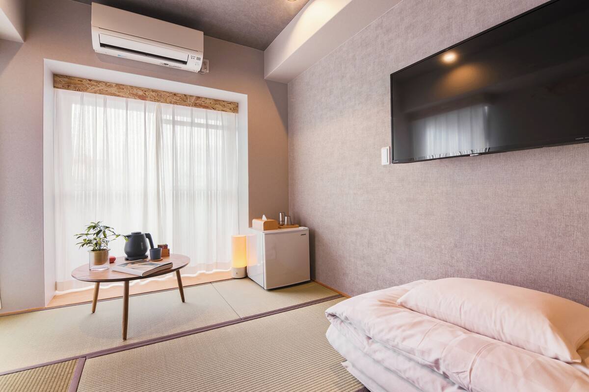 Ein Hotelzimmer mit Tatami-Boden, einem Bett mit Kissen, einem kleinen Tisch mit Pflanze und Wasserkocher, einem Kühlschrank, einem an der Wand montierten Fernseher und einem Fenster mit durchsichtigen Vorhängen.