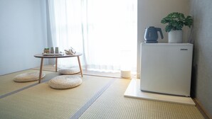 A minimalist Japanese-style room with tatami flooring, a small table with tea set, a refrigerator, and a potted plant. - Wayfarer Hotel Kyoto Shijo (Kyoto)