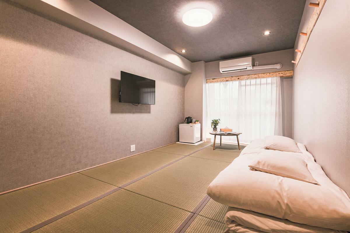 Ein modernes Hotelzimmer mit Tatami-Boden, einem Flachbildfernseher, einem kleinen Tisch mit Pflanze und einem Bett mit Kissen.