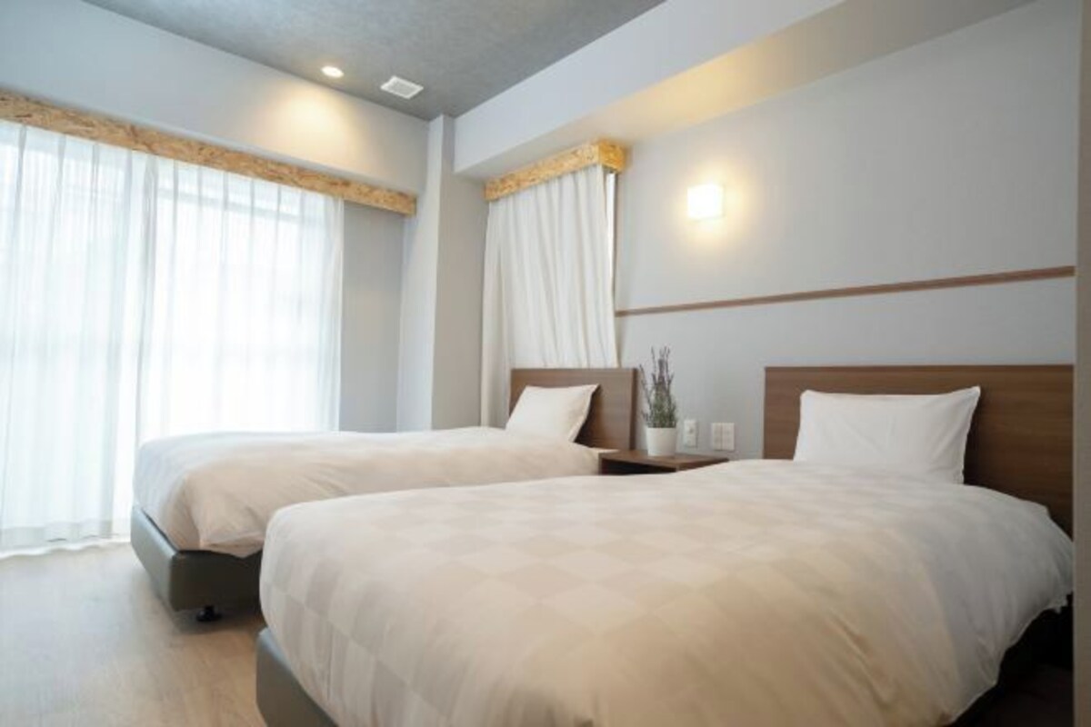 A hotel room with two beds, a wooden headboard, a bedside table with a lamp, and a window with sheer curtains.
