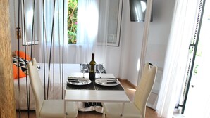 Un coin repas avec une table blanche dressée pour deux personnes, une bouteille de vin et deux verres. Il y a deux chaises avec un coussin de couleur claire. La pièce est dotée d’un plancher en bois et d’une fenêtre avec des rideaux.