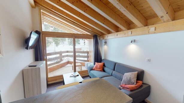 A cozy room with a wooden ceiling, a grey sofa with cushions, a small table, and a TV mounted on the wall.