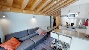 A modern kitchen with a microwave, sink, and stainless steel appliances. There is a grey sofa with orange cushions and a small white table.