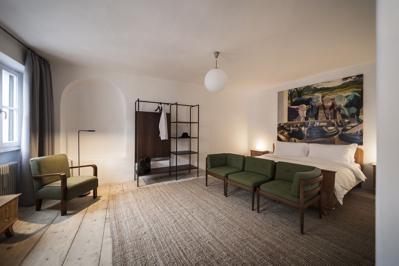 A modern room with a green sofa, a wooden chair, a bed with white linens, a black metal wardrobe, and a painting of cows on the wall.