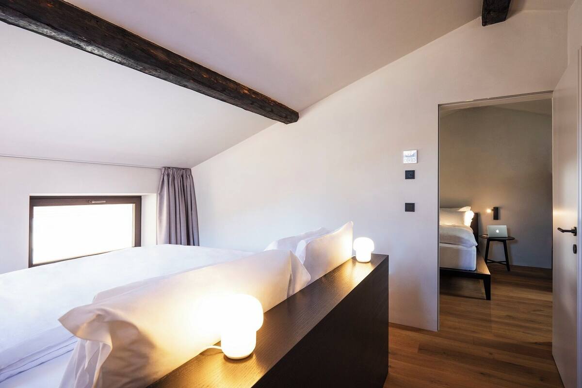 A modern hotel room with a large bed, bedside lamps, a wooden headboard, and a view of another room.