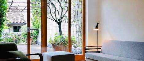 A room with a wooden ceiling, a sofa, a chair, a floor lamp, and large windows with a view of trees and a building outside.