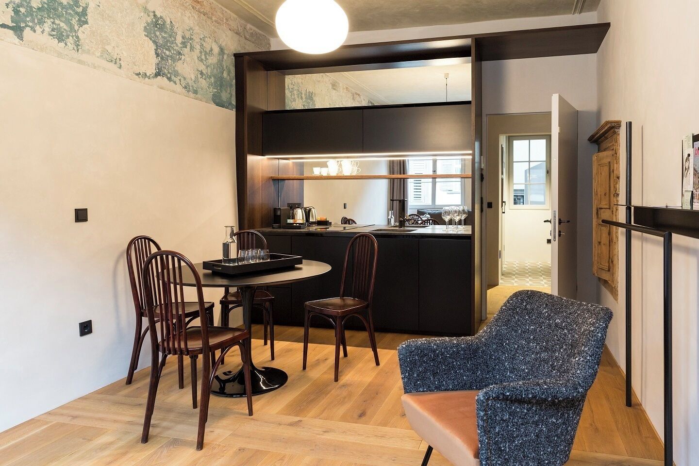Une cuisine compacte dotée d’appareils électroménagers modernes, un coin repas avec des chaises en bois et un petit espace salon avec un fauteuil à motifs.