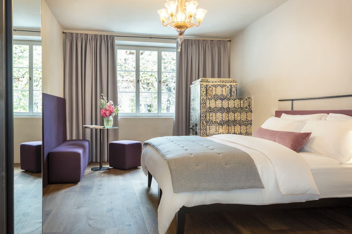 A hotel room with a large bed, a small table with a vase, two purple armchairs, and a decorative cabinet.
