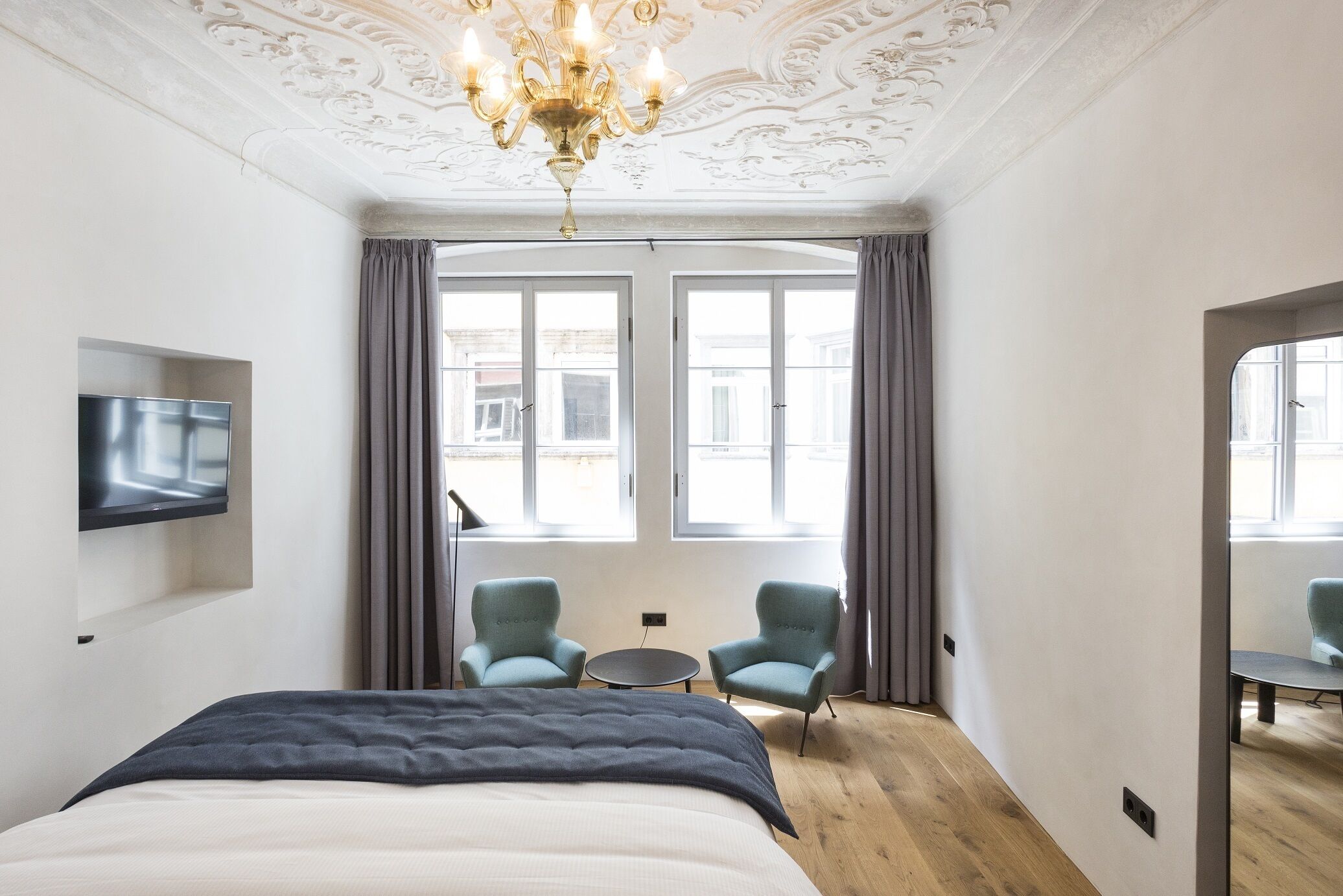 Une chambre d’hôtel avec un grand lit, un téléviseur fixé au mur, deux fauteuils, une petite table et un lustre.