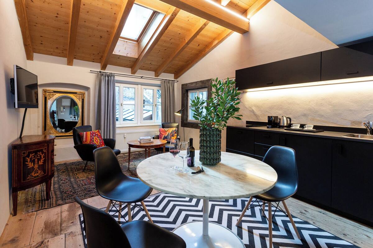 A modern kitchen with a dining area, wooden ceiling beams, and a large window.